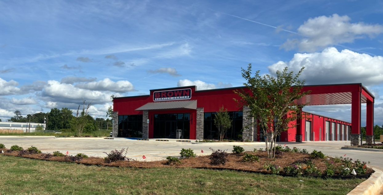 Red commercial building with a black awning and sign, set beside a parking lot and landscaped grass under a blue sky