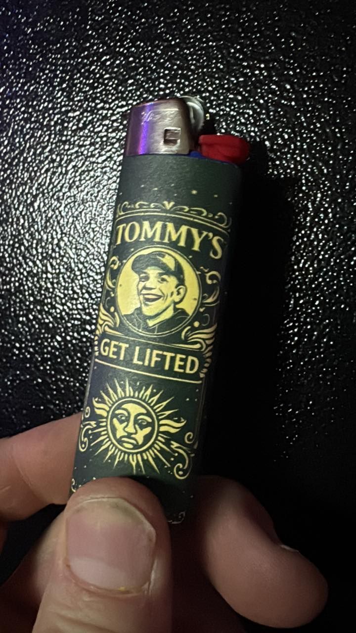 Hand holding a black and gold Tommy’s “Get Lifted” lighter against a dark background