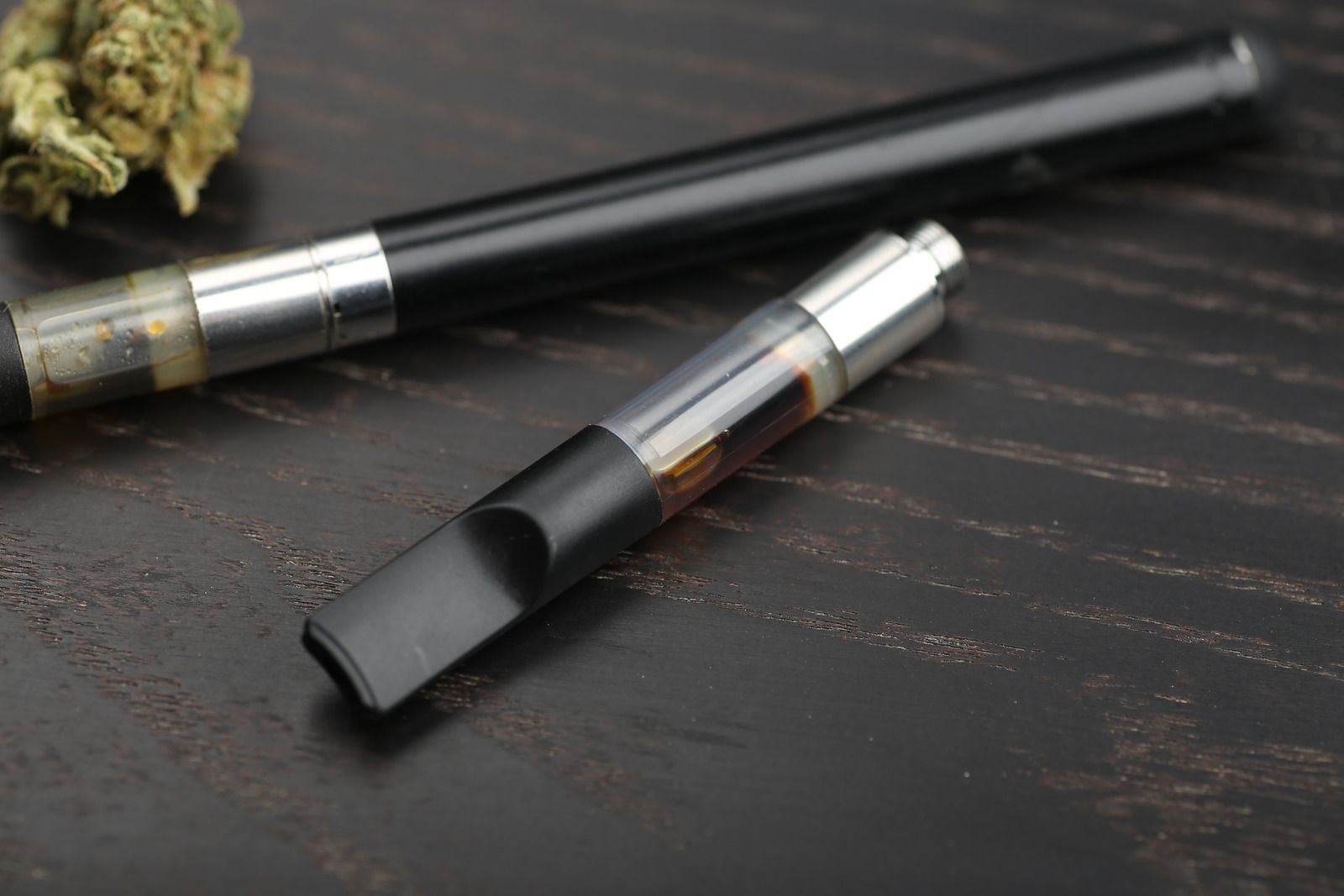 Two black vape pens on a dark wood surface, one with a clear tank and mouthpiece in the foreground