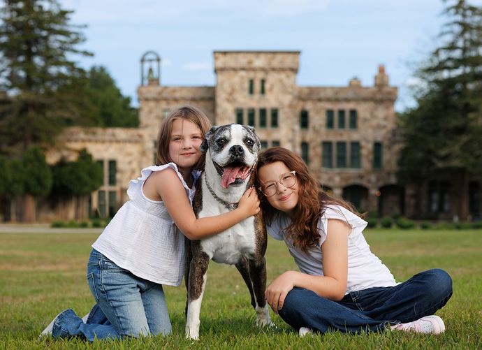Two girls hugging a dog in front of a stone building on a grassy lawn.