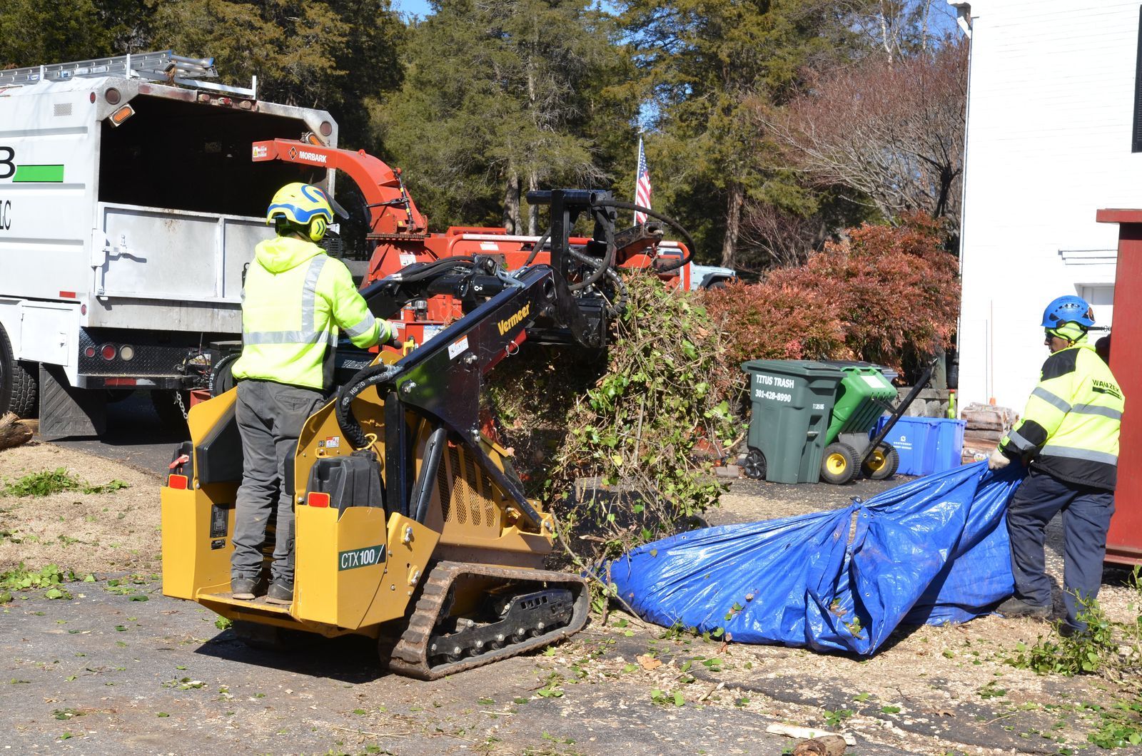 Two workers loading brush into a wood chipper using a skid steer, parked near a building.