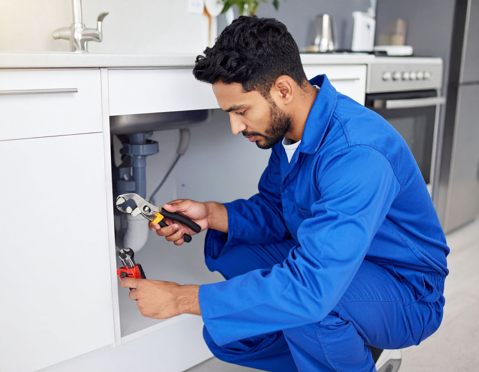 A person in blue coveralls kneels under a kitchen sink, using a tool to repair the plumbing pipes.