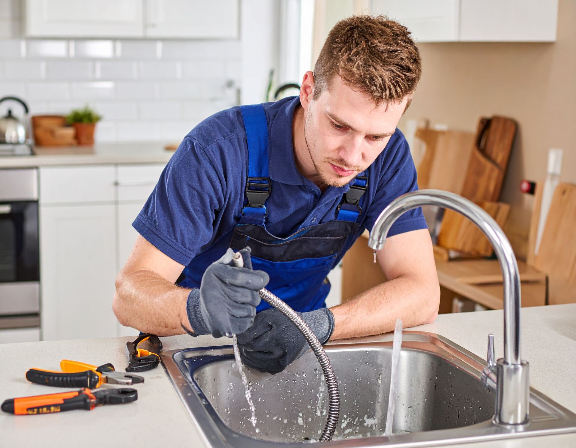 A plumber in blue overalls and gloves working on a kitchen sink faucet with water spraying into the stainless sink.