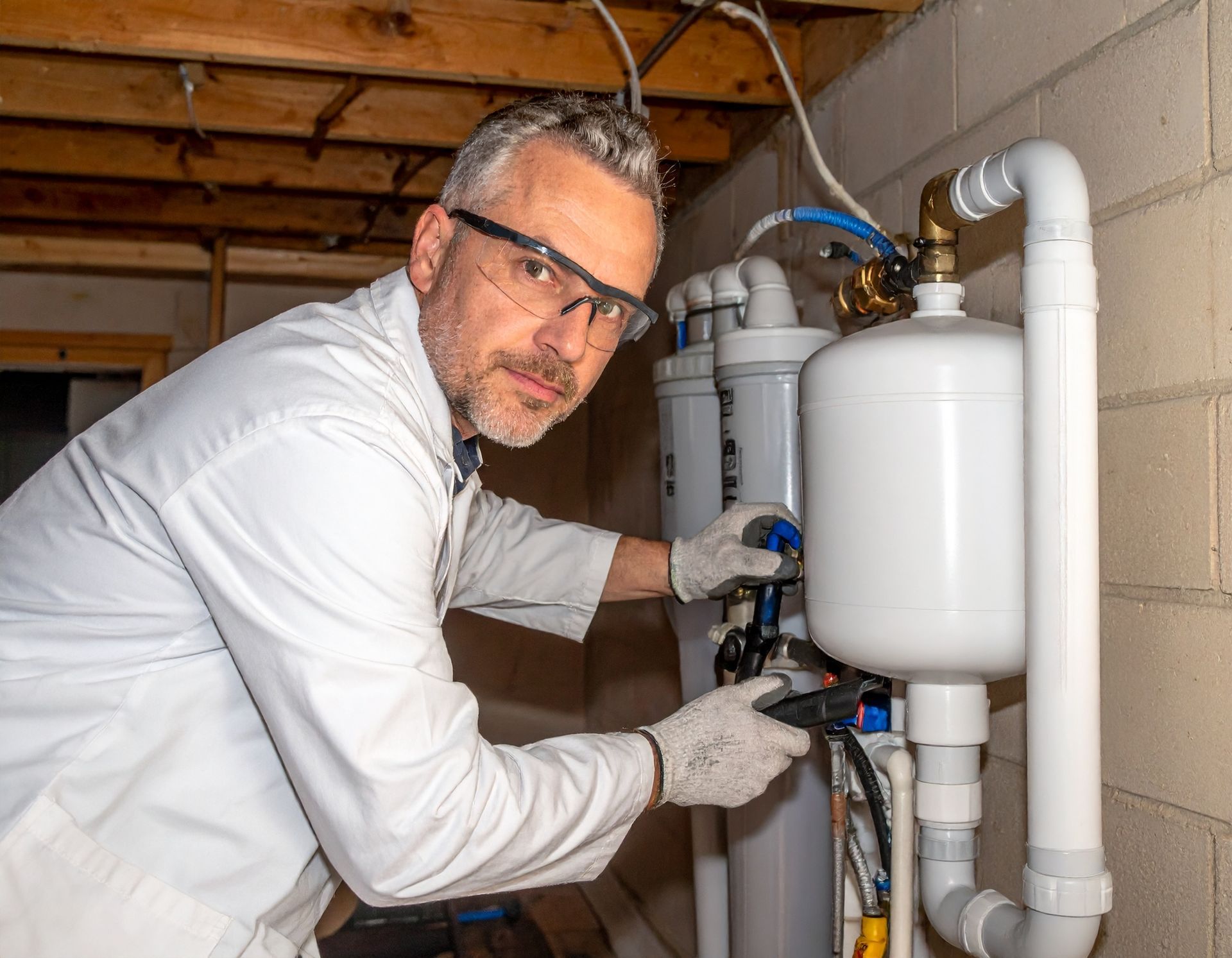 A professional in a lab coat and safety glasses wearing work gloves while servicing a residential water filtration system.