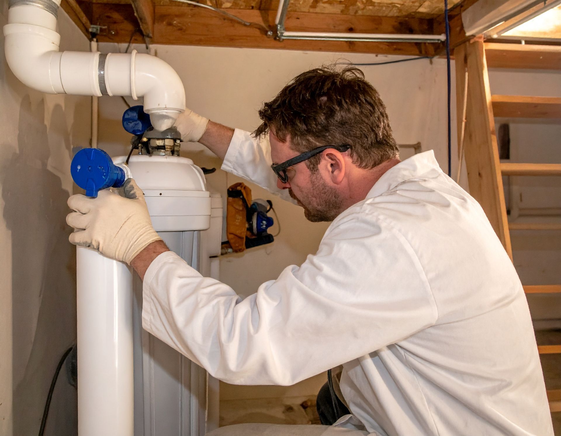 A person in a white lab coat and safety glasses services a white industrial water filtration unit in a basement.