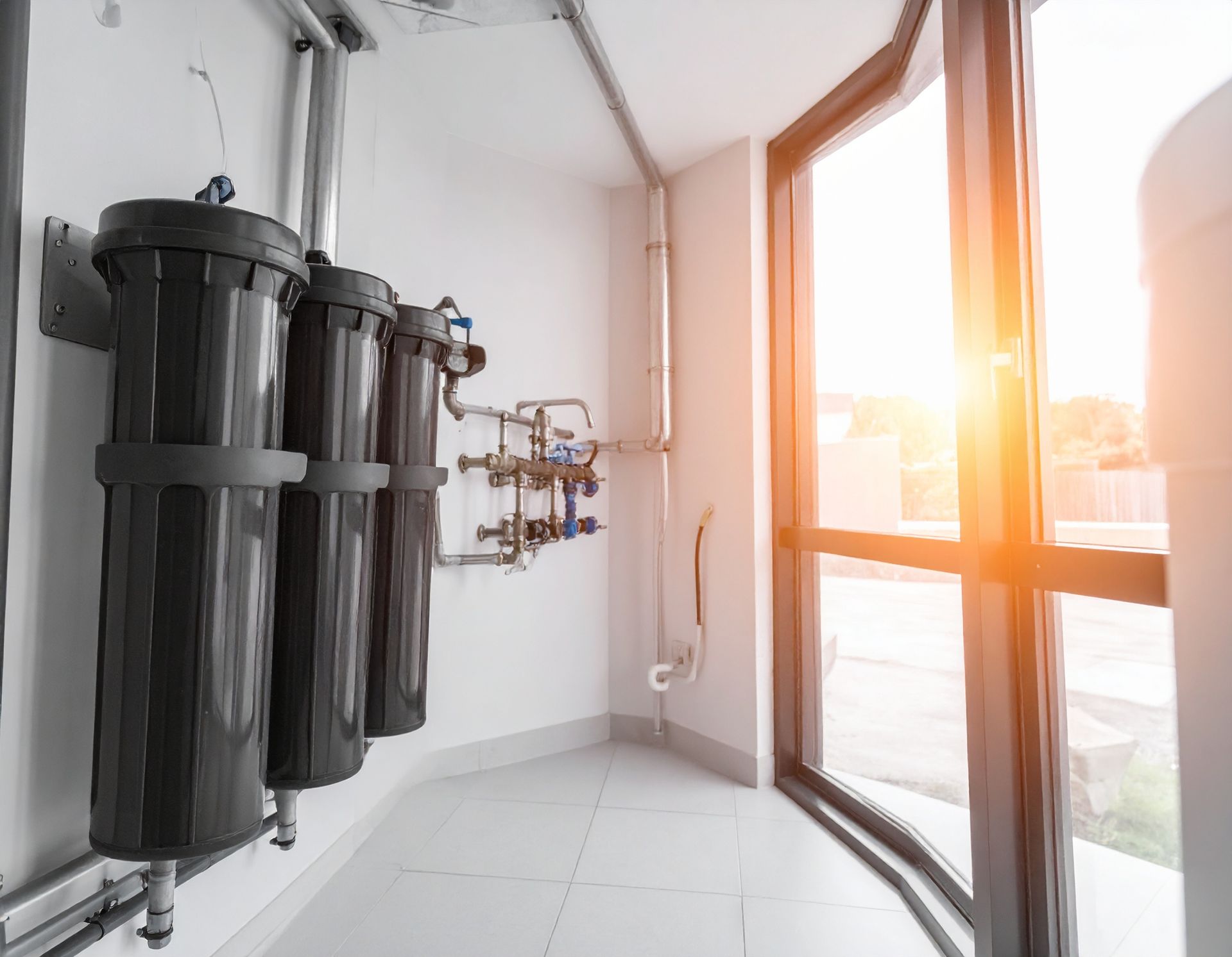 Three black water filter cylinders mounted on a white wall with metal plumbing pipes, near a large window.
