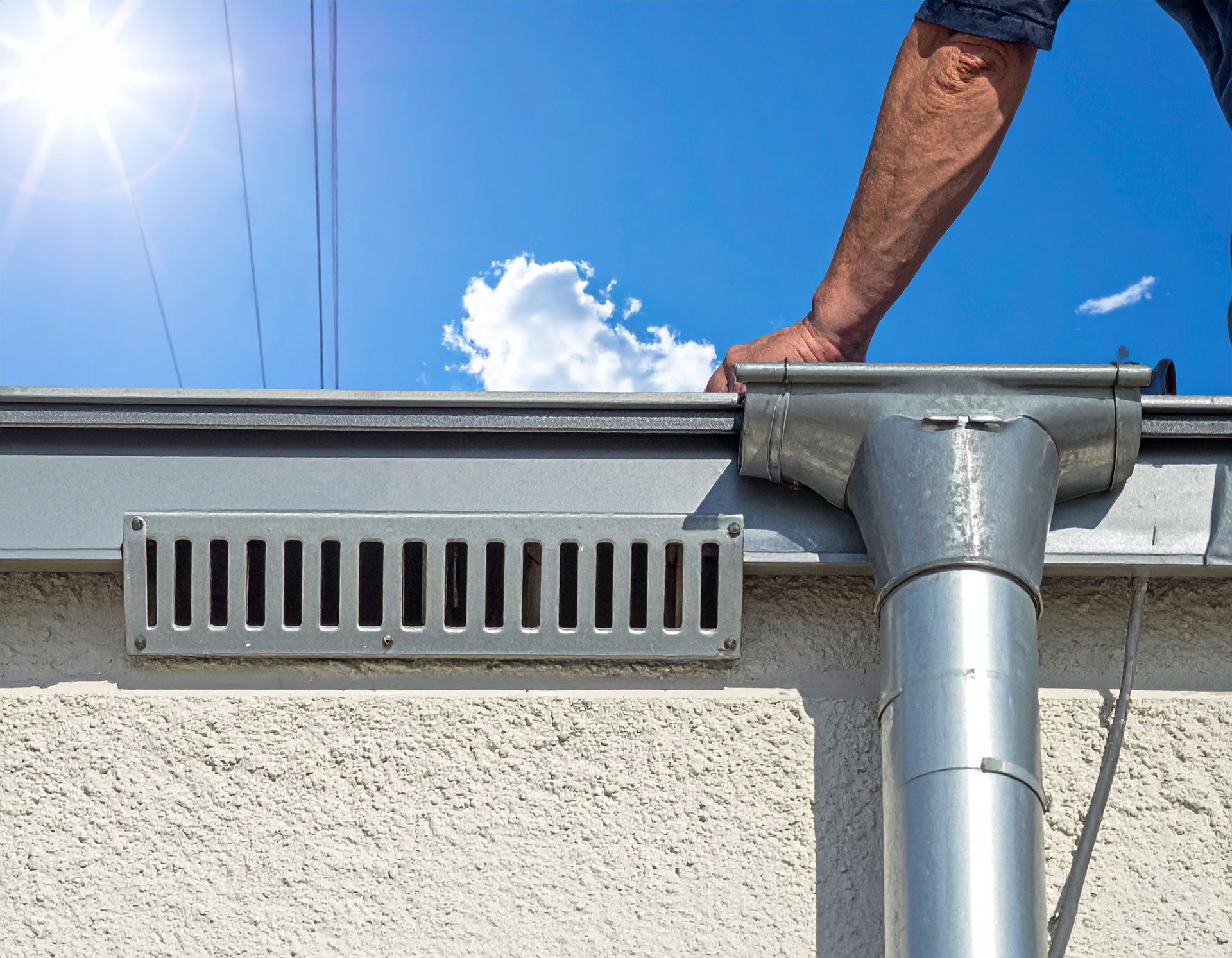 A worker cleans a metal gutter on the side of a building, located next to a rectangular wall vent against a blue sky.