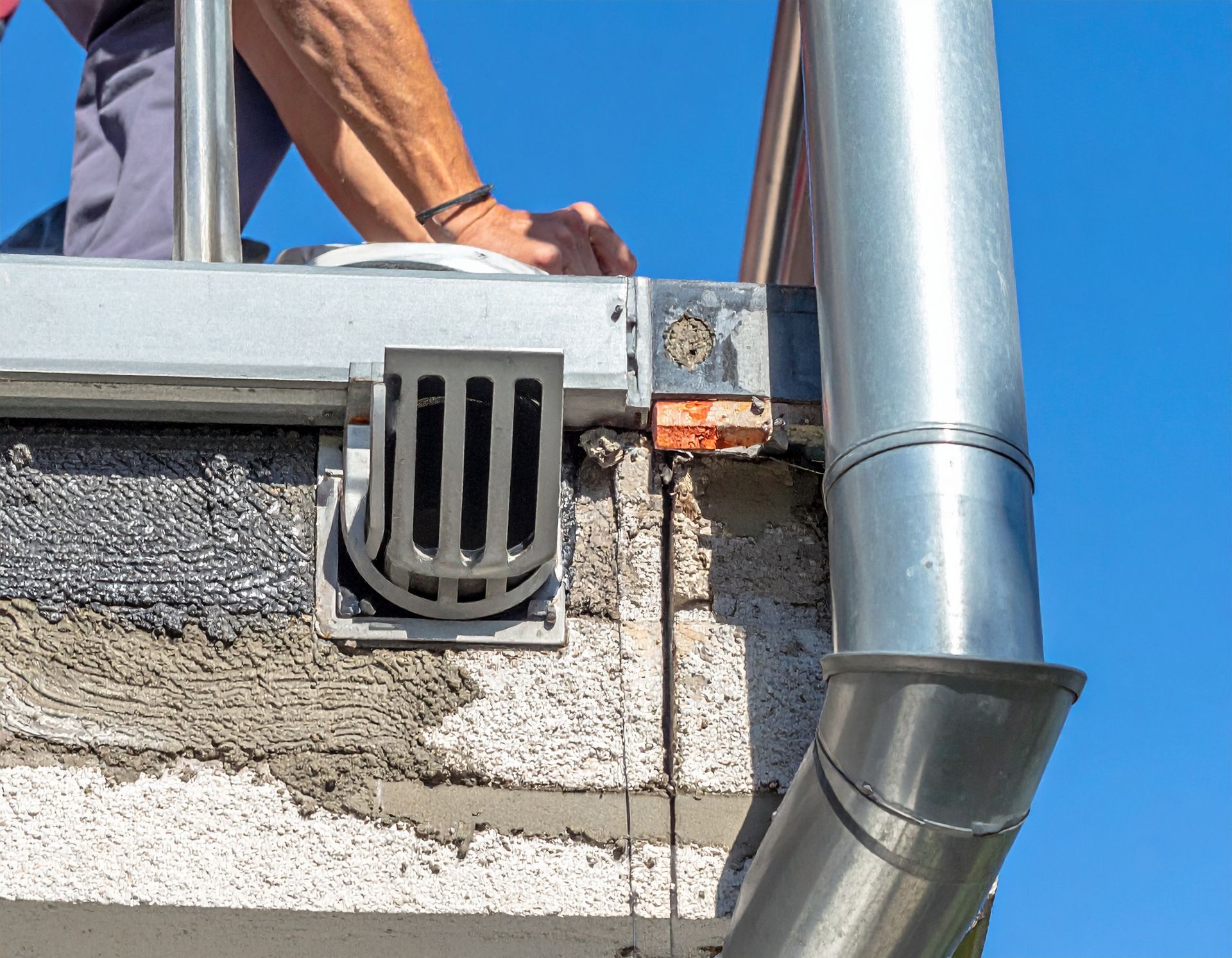 A person works on a flat roof edge featuring a metal drainage grate and an attached vertical gutter pipe.