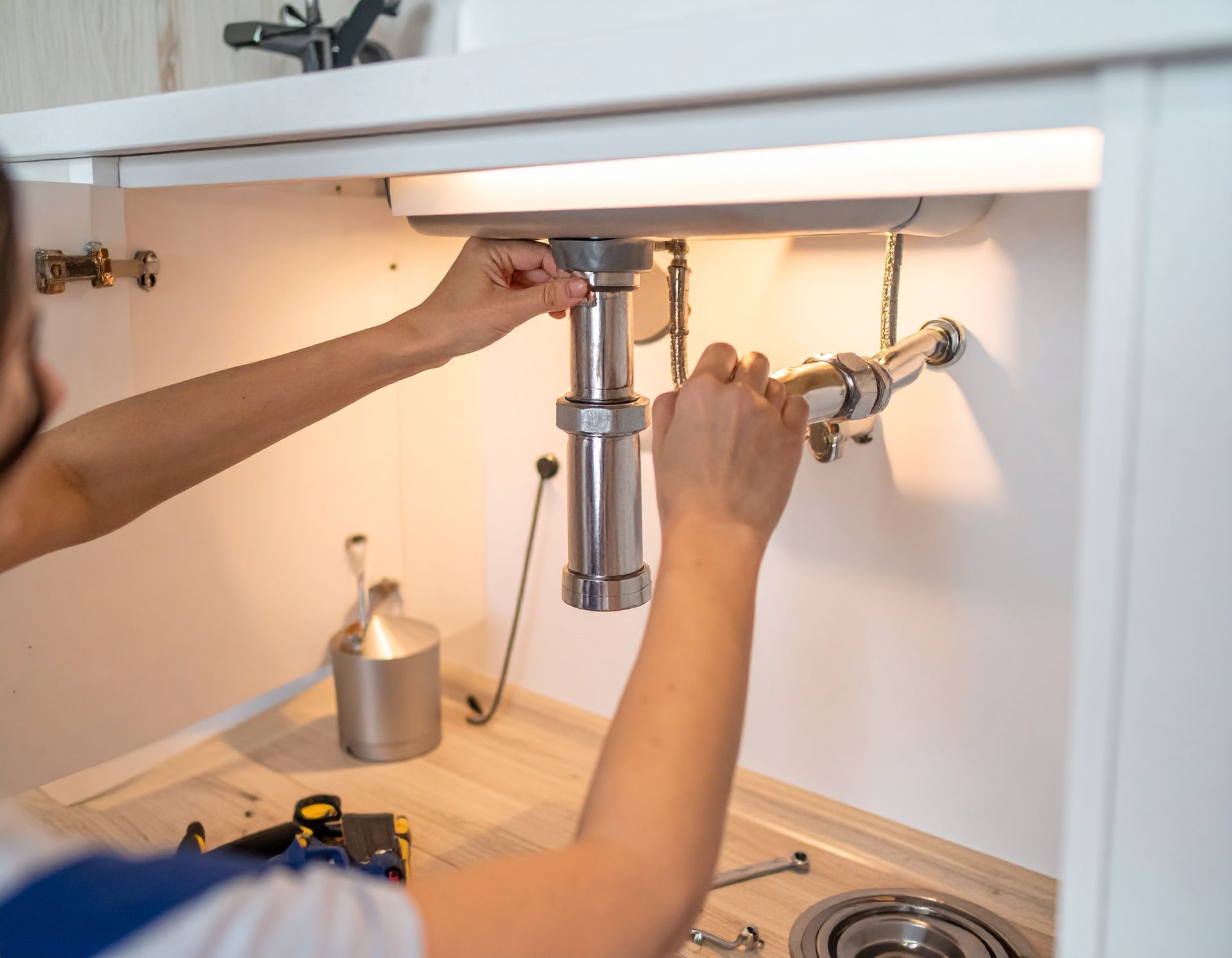 A person repairing the metal plumbing pipes under a white kitchen sink.