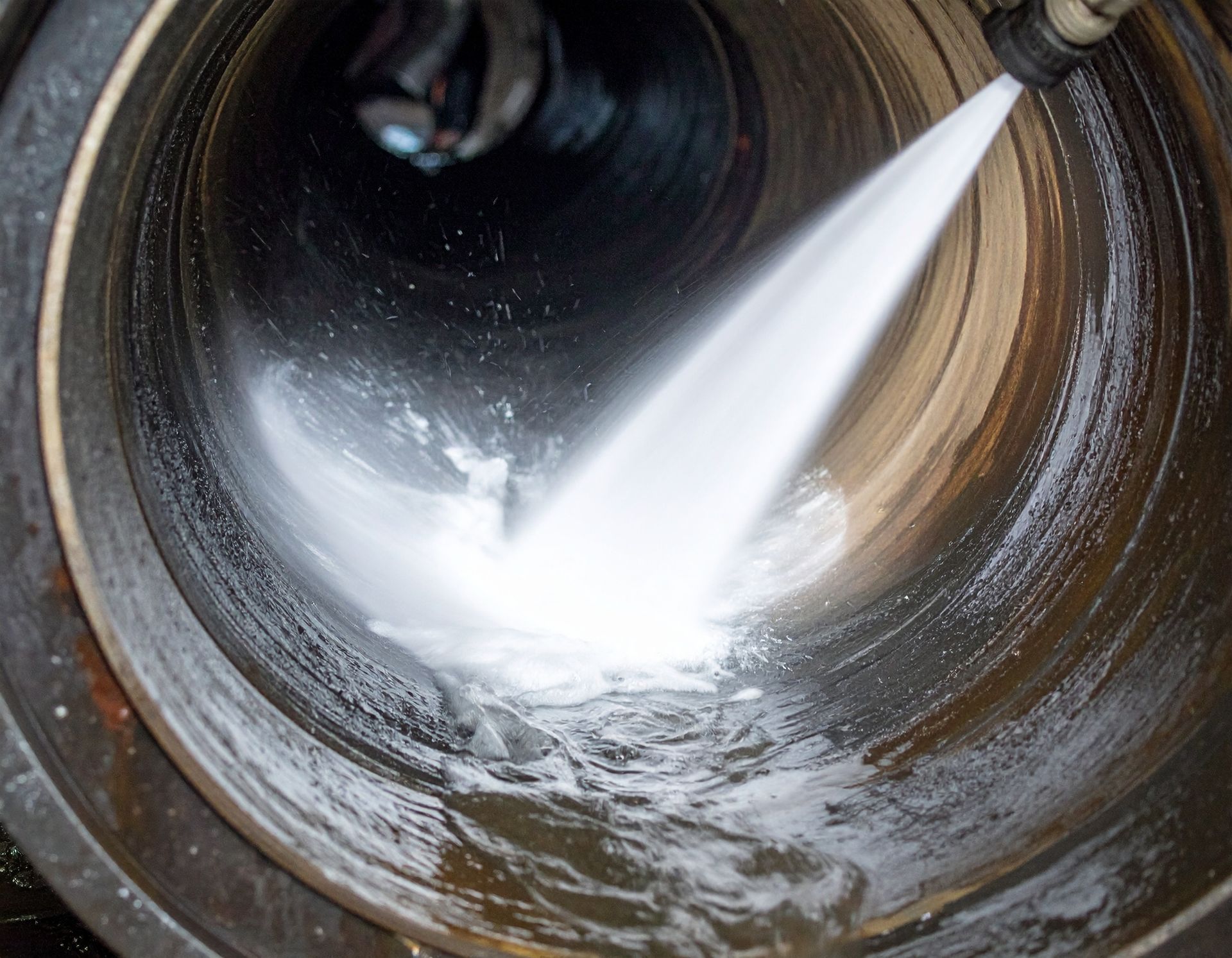 A high-pressure water nozzle cleaning the interior of a dark, cylindrical pipe, revealing a clean, metallic surface.