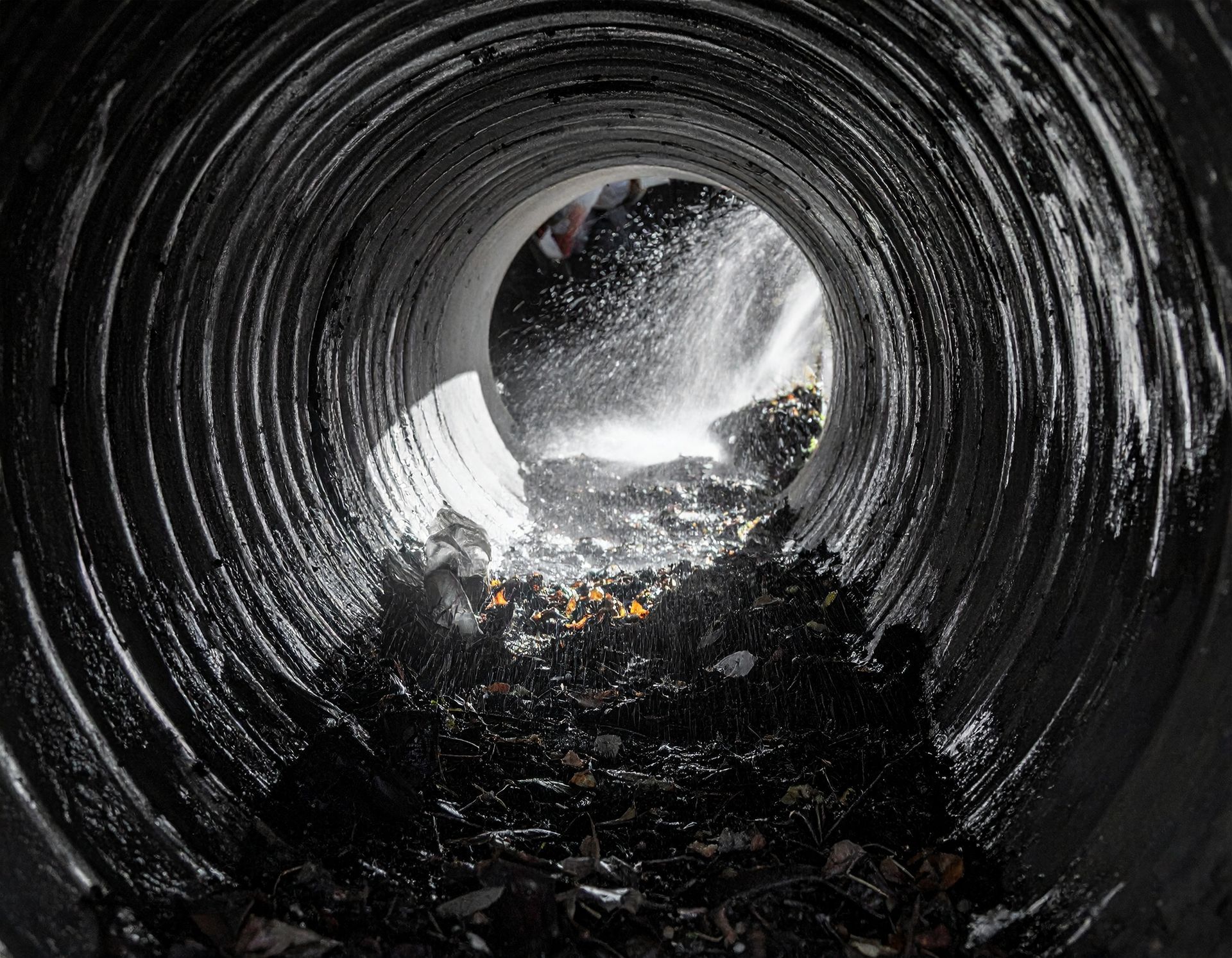 A view from inside a corrugated pipe, looking toward a bright opening where water splashes over debris and rocks.