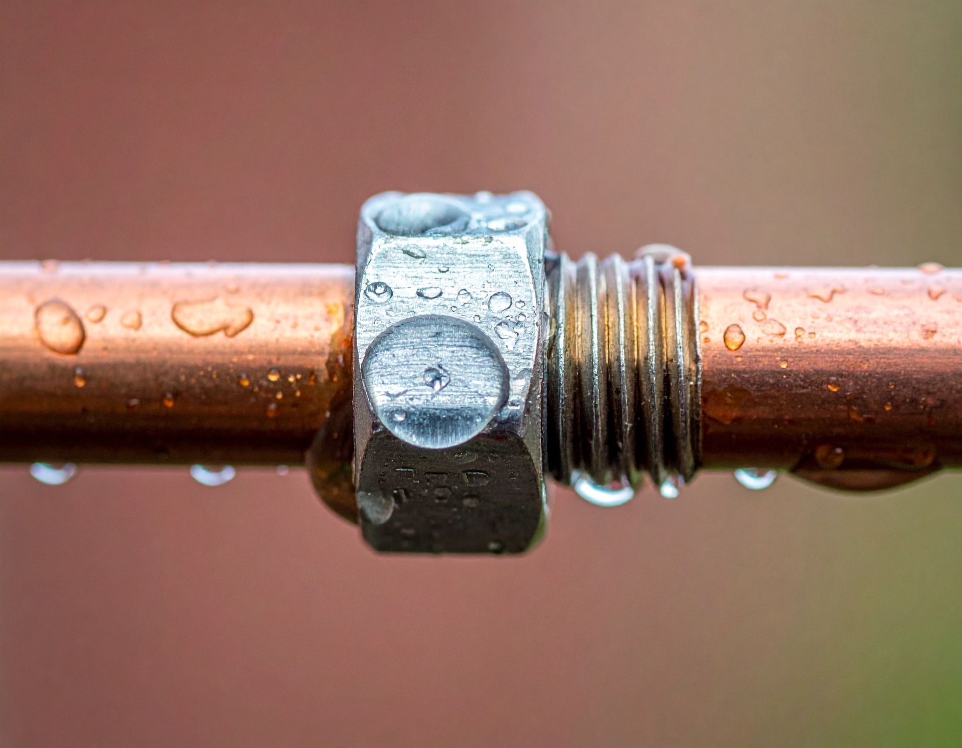 A close-up view of a metal nut threaded onto a copper pipe, with water droplets on the metallic surfaces.