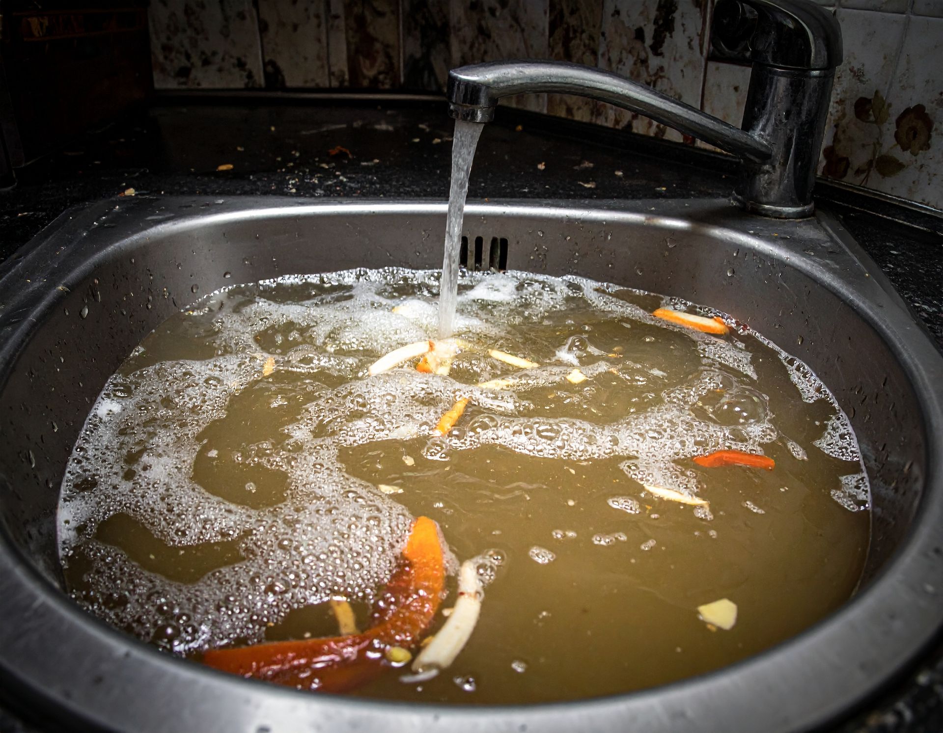 A clogged kitchen sink filled with murky, soapy water and food scraps, with water running from the faucet.