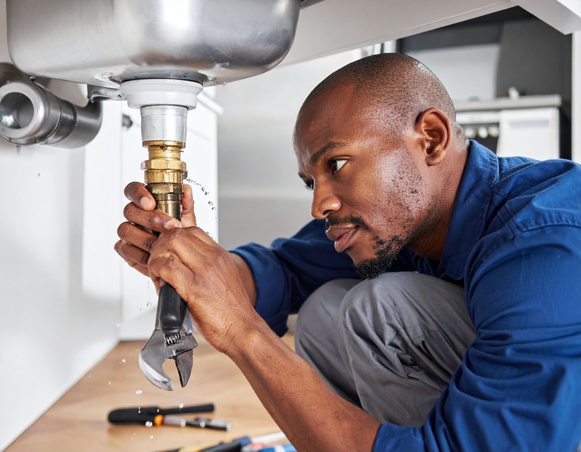 A person in a blue shirt uses an adjustable wrench to repair a leaking metal pipe underneath a kitchen sink.