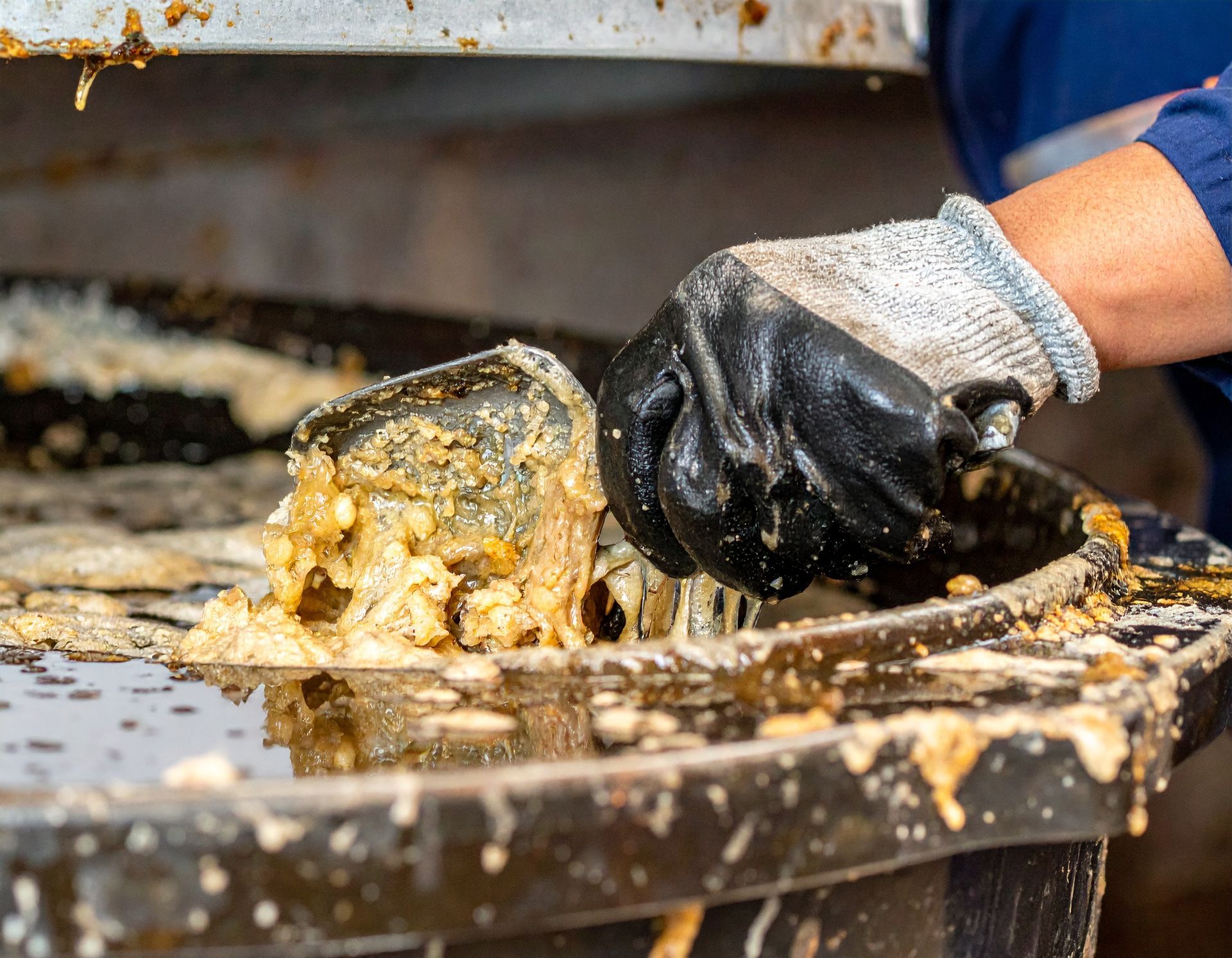 A gloved hand uses a tool to stir a thick, brownish substance in a large, dark industrial vat.