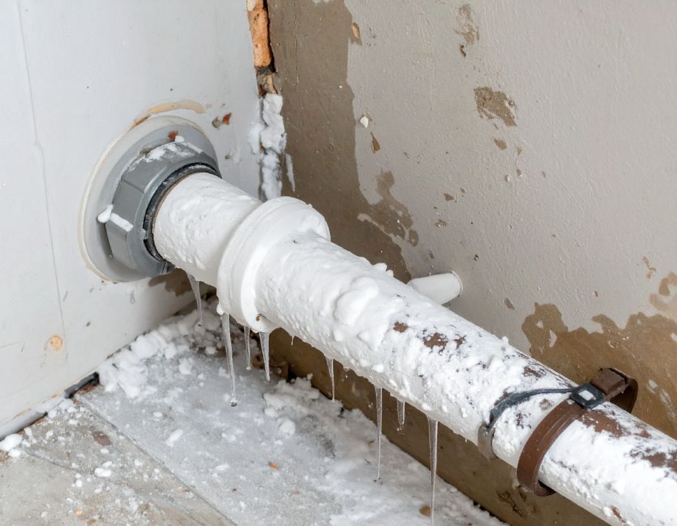 Frozen pipe, covered in ice, connected to a white appliance, inside a damaged wall.