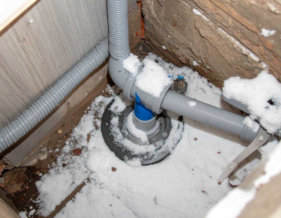 Snow-covered plumbing pipes in a recessed area, with a corrugated grey hose, connecting to a blue-gray junction.