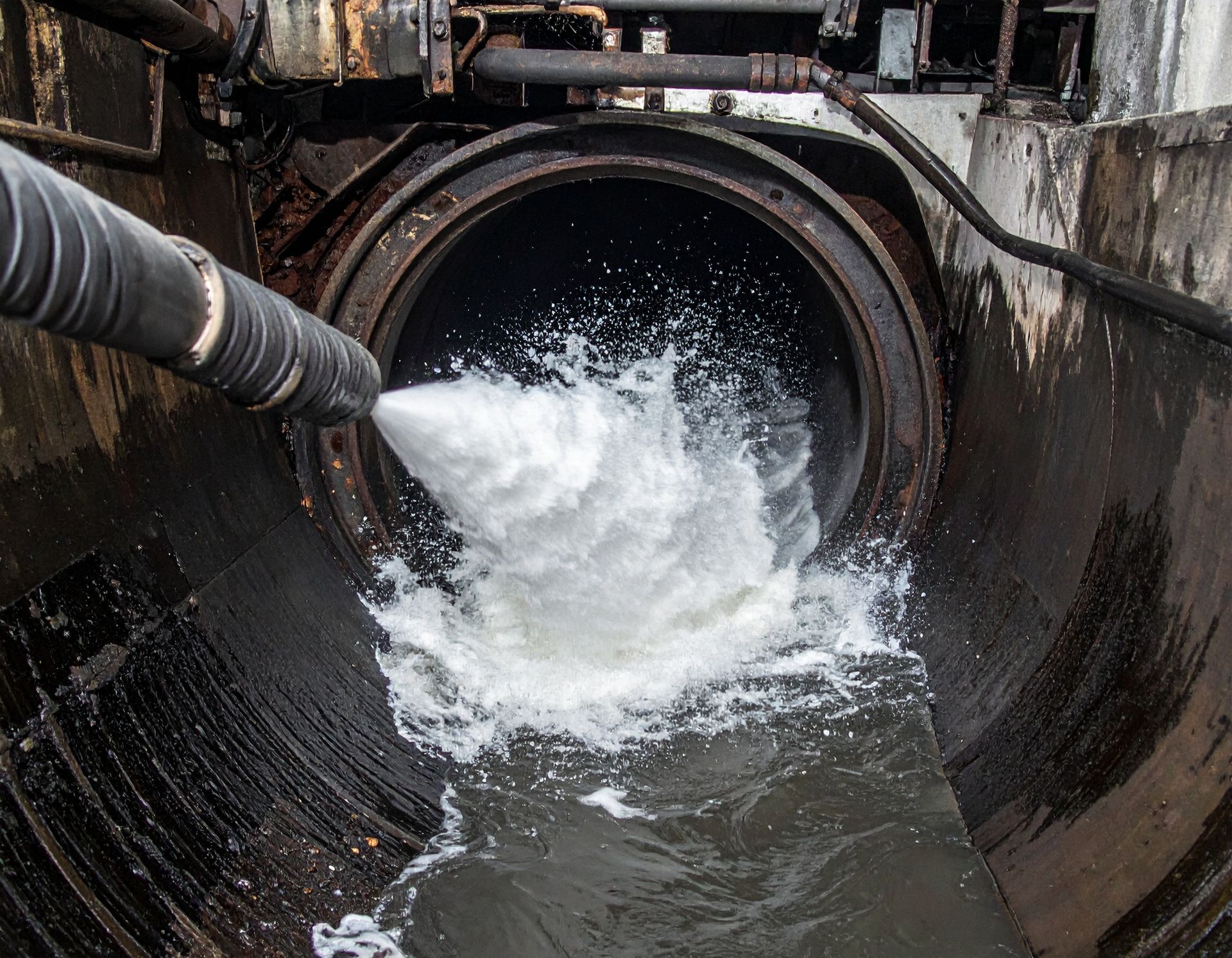 A pressurized hose sprays a white liquid into a large, dark circular pipe at an industrial facility.