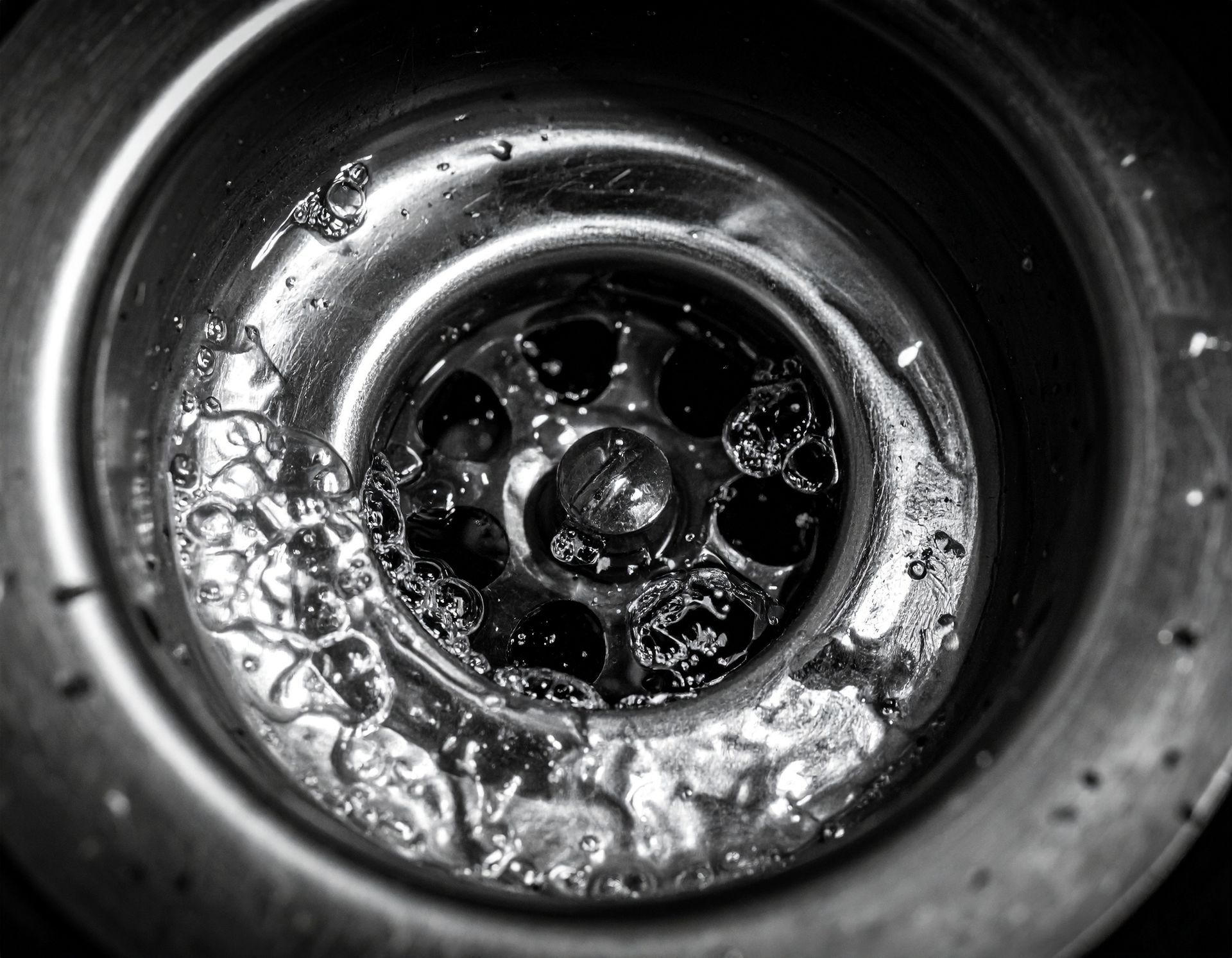 A close-up, black-and-white view of a metal kitchen sink drain containing standing water and small bubbles.