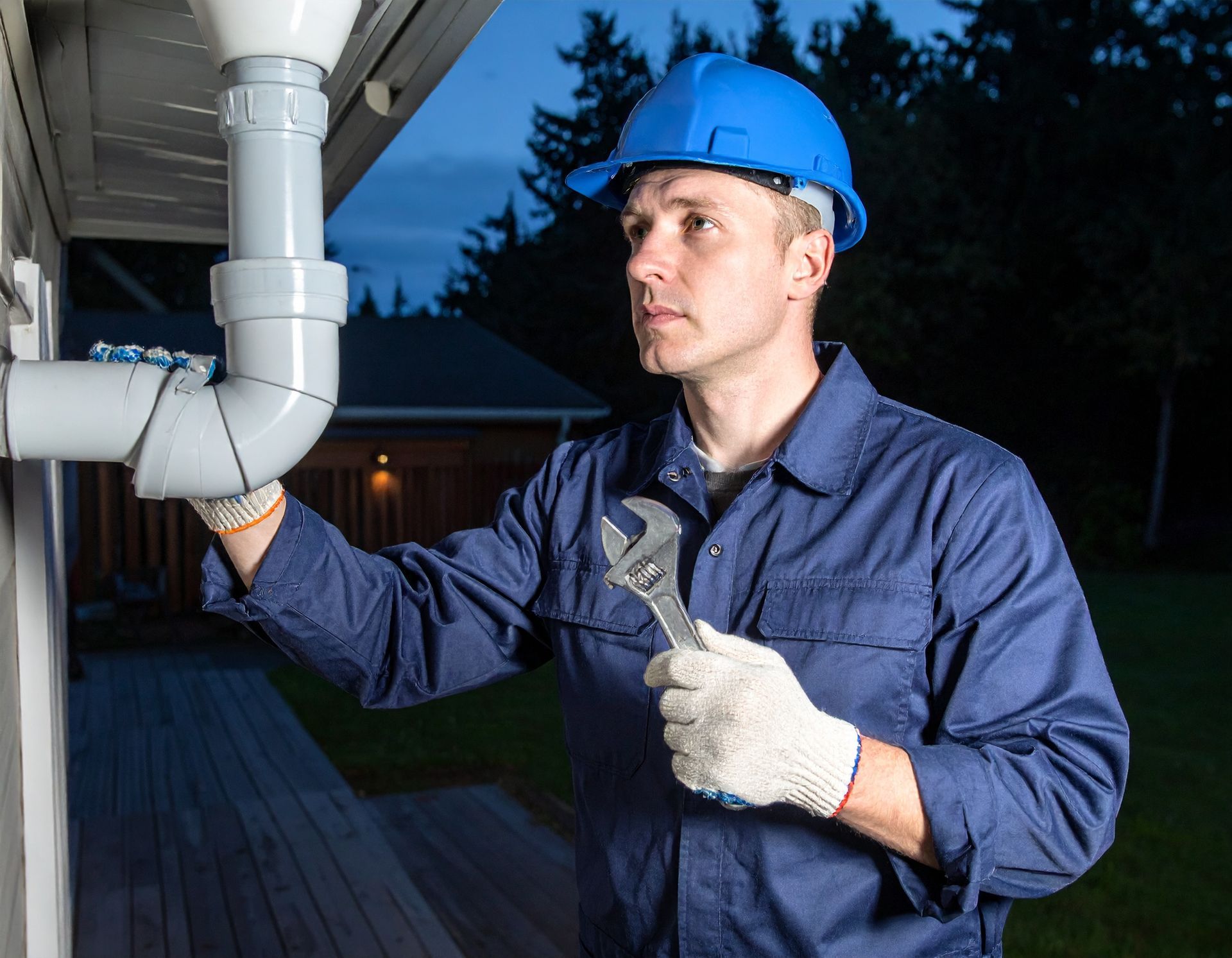 A person in a blue hard hat and uniform holds an adjustable wrench while checking a grey pipe on an exterior wall.