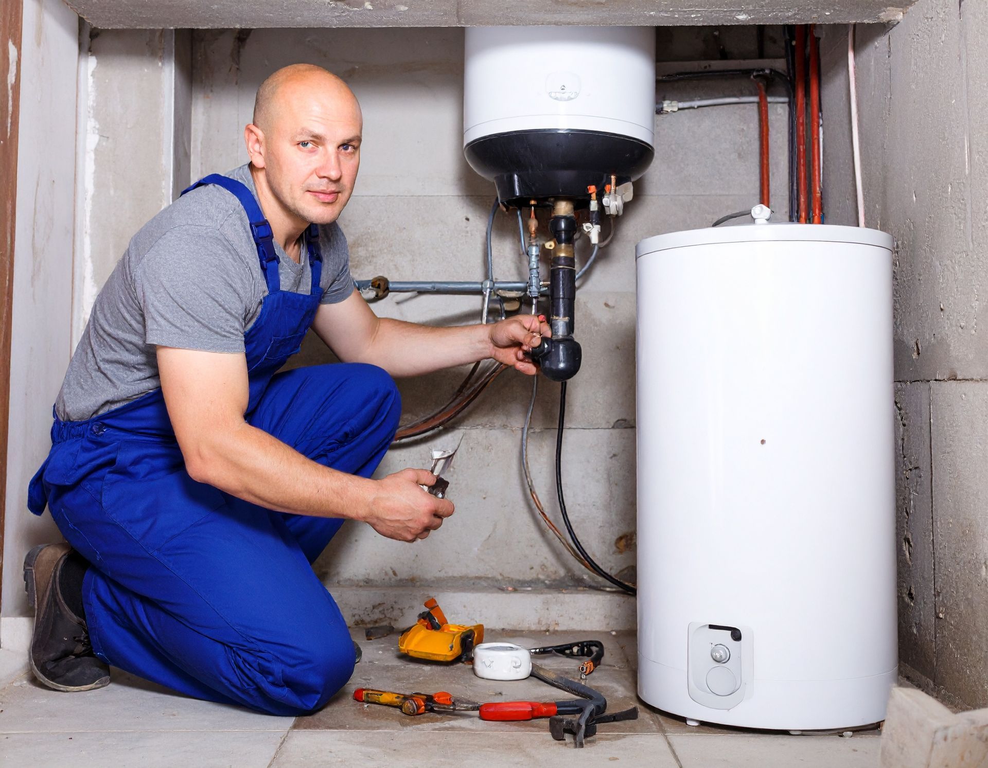 A technician in blue coveralls kneels on a concrete floor, repairing a wall-mounted water heater with various tools.