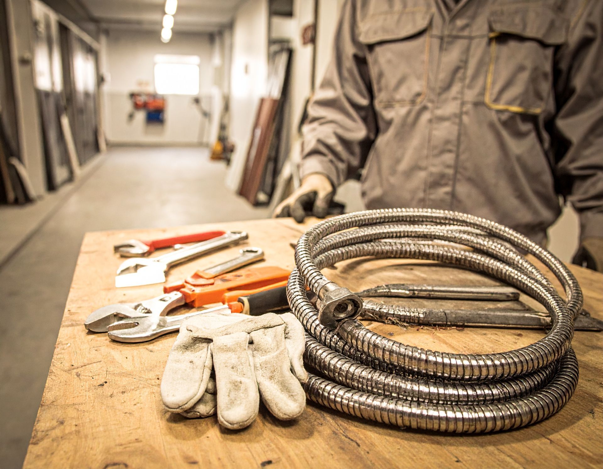 A worker standing at a workbench with tools and a coiled flexible metal hose in a workshop.