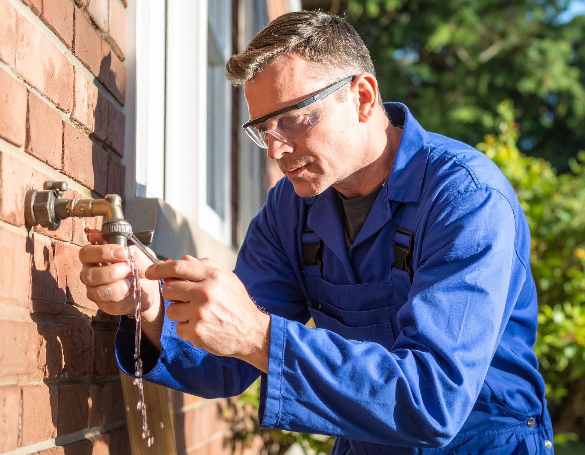A person in blue work coveralls and safety glasses repairs an outdoor water faucet attached to a brick wall.