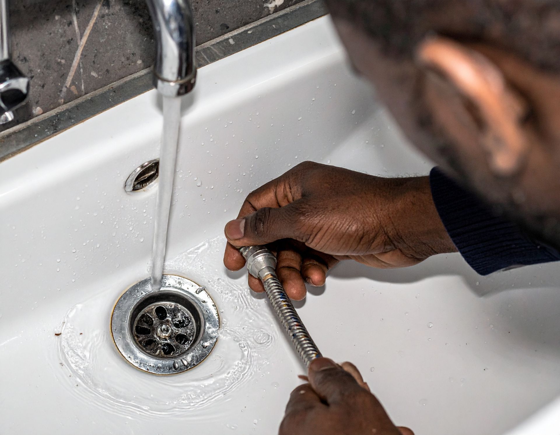 A person holds a metal plumbing component near a white sink basin while water flows from a faucet into the drain.