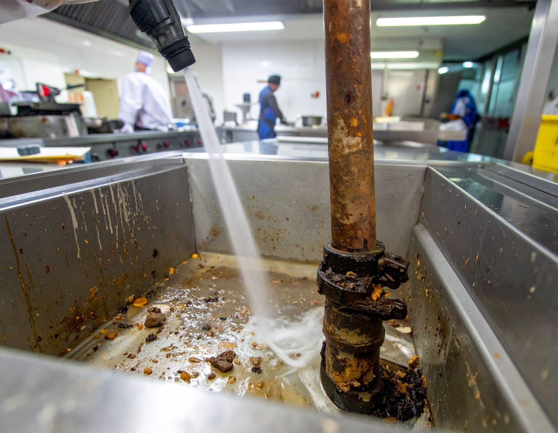 A high-pressure spray nozzle cleans a dirty, rusted pipe inside a stainless steel basin in a commercial kitchen.