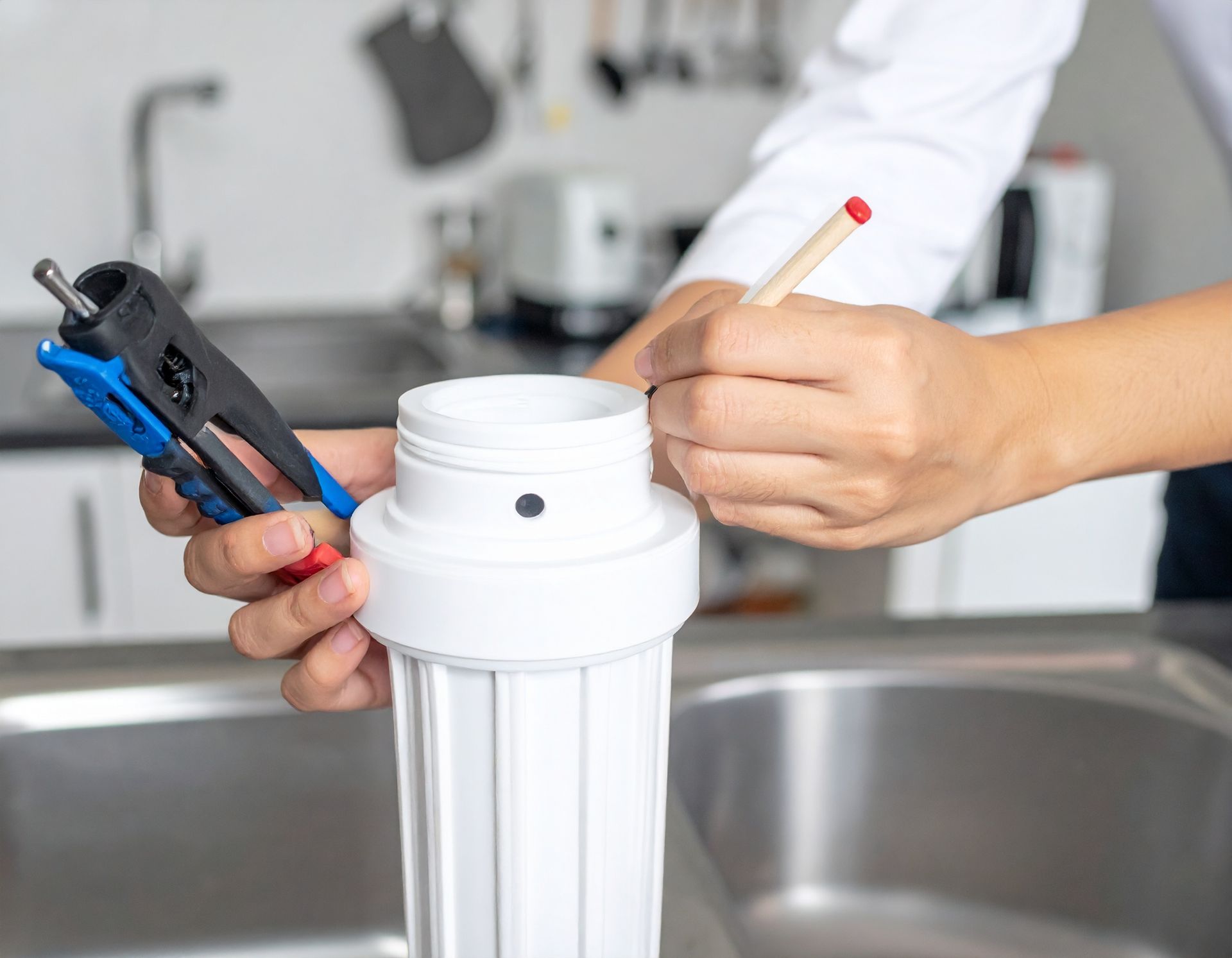 A person holds a water filter housing over a kitchen sink while using a pen and a specialized wrench to work on it.