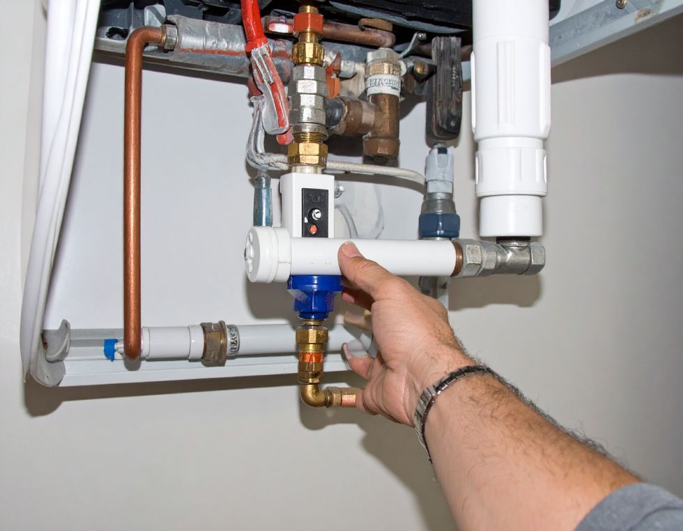 Hand touching a water filter connected to a boiler system. Copper pipes and white components are visible.