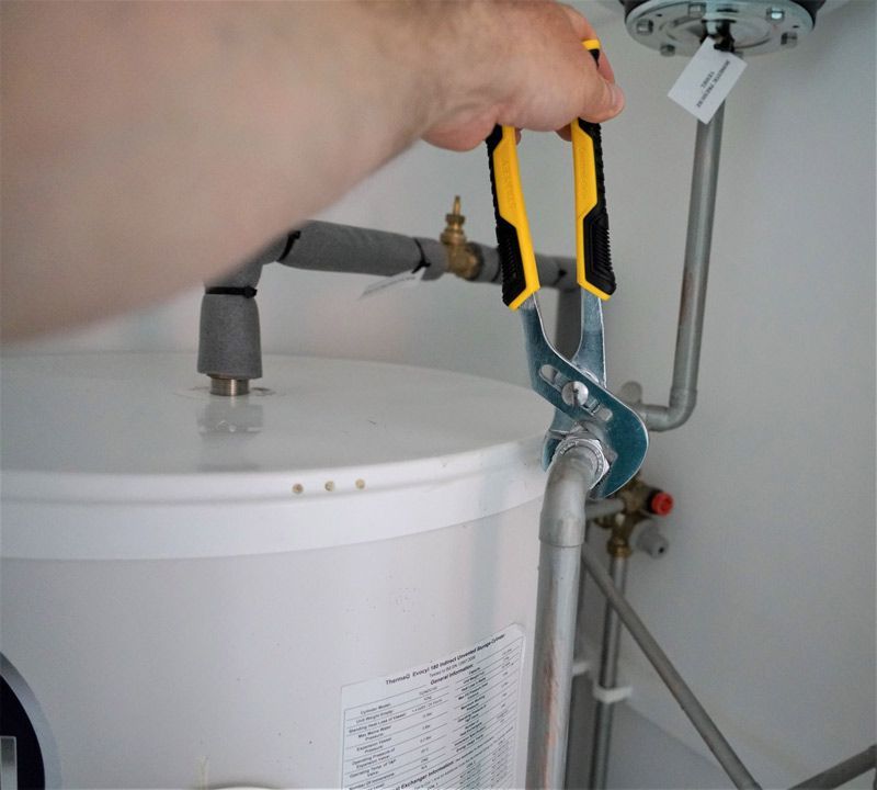 A hand uses yellow-handled tongue-and-groove pliers to tighten a metal plumbing fitting on a water heater.