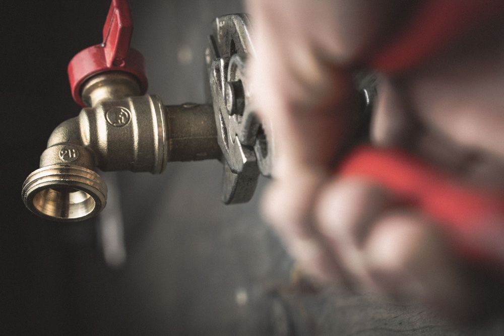 A close-up view of hands using pliers to tighten a brass faucet with a red handle against a dark background.