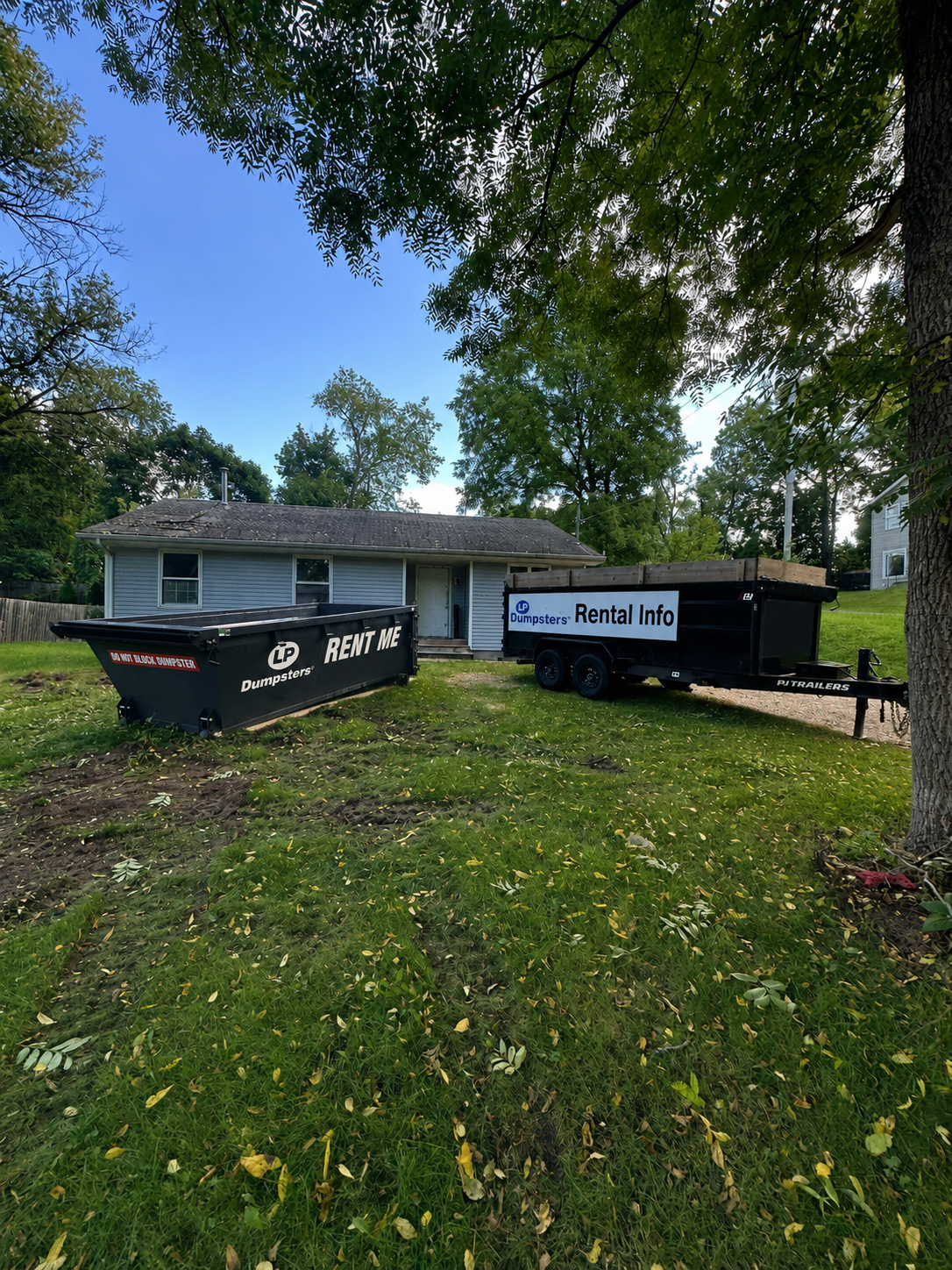 Two black dumpster bins in a grassy yard beside a white house under trees.
