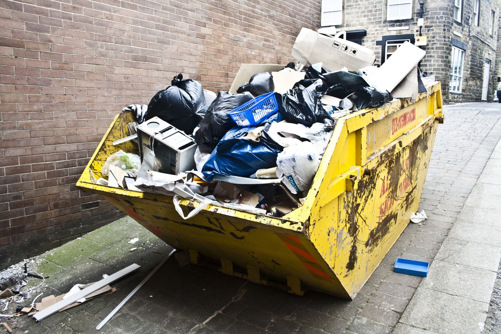 Overflowing yellow dumpster full of trash in a narrow alley beside a brick wall