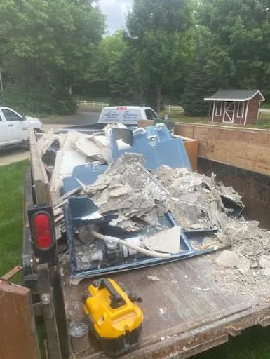 Truck bed filled with broken debris and a yellow tool in a driveway, with parked cars and trees behind.