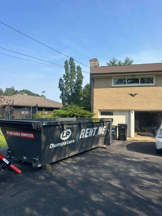 Large black dumpster labeled “RENT ME” in a residential driveway beside a tan house.
