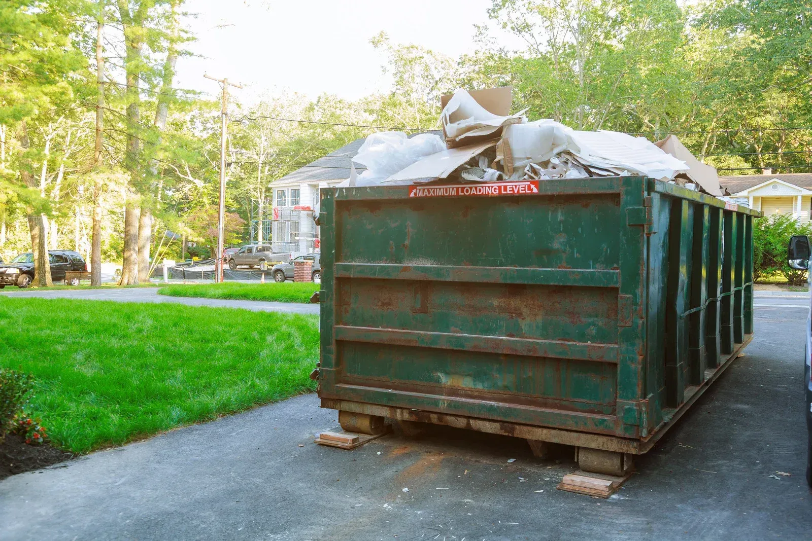 Green dumpster filled with trash on a residential street beside a grassy yard