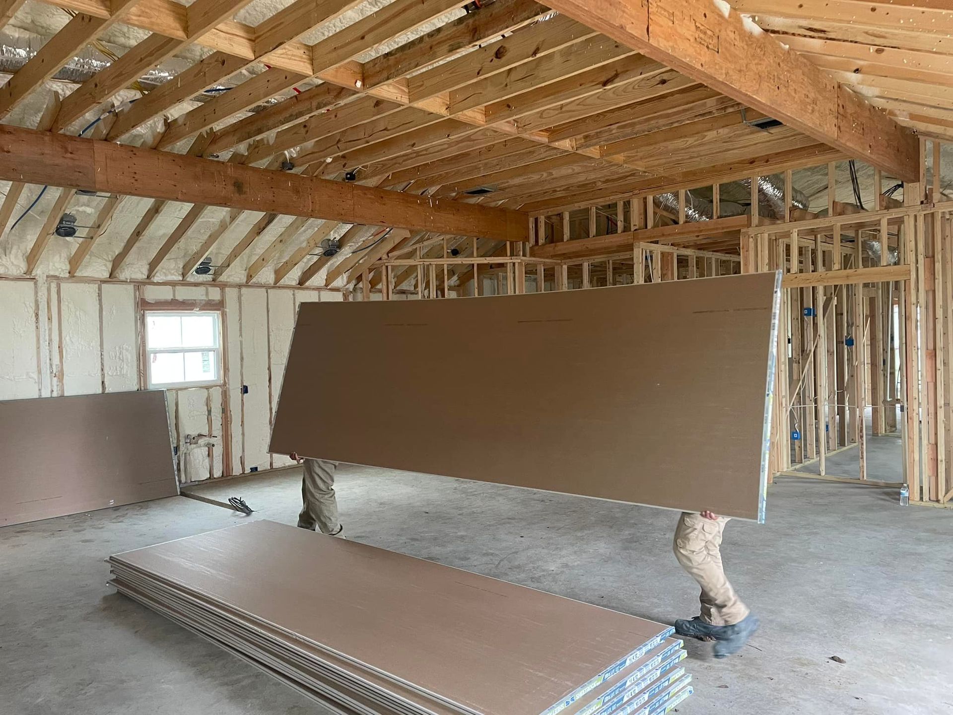 A man is carrying a large piece of drywall in a room under construction.
