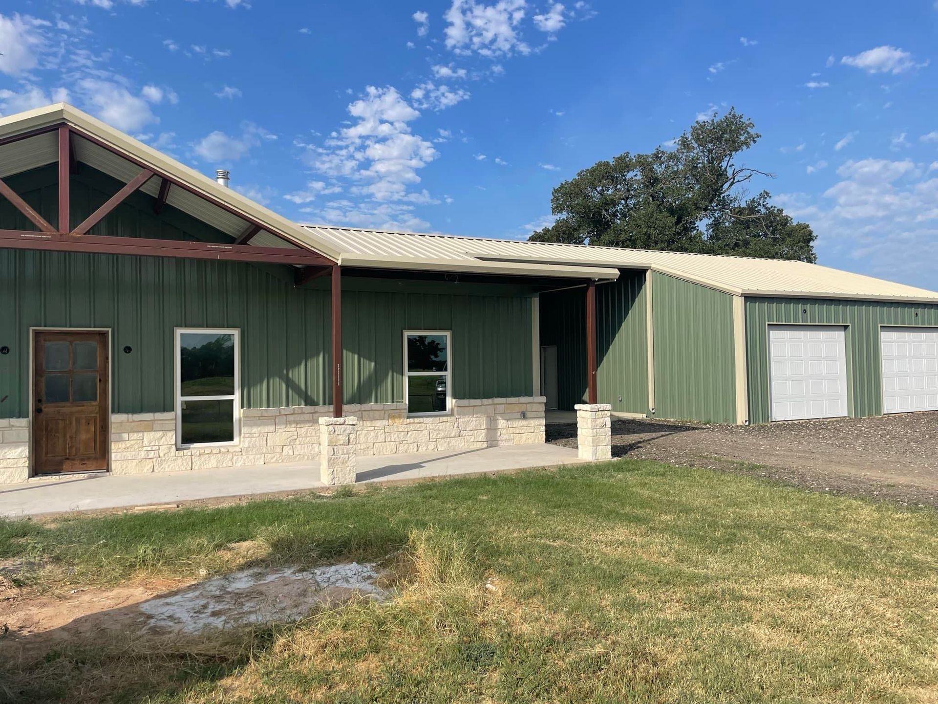 A green metal building with a porch and garage doors is sitting on top of a lush green field.