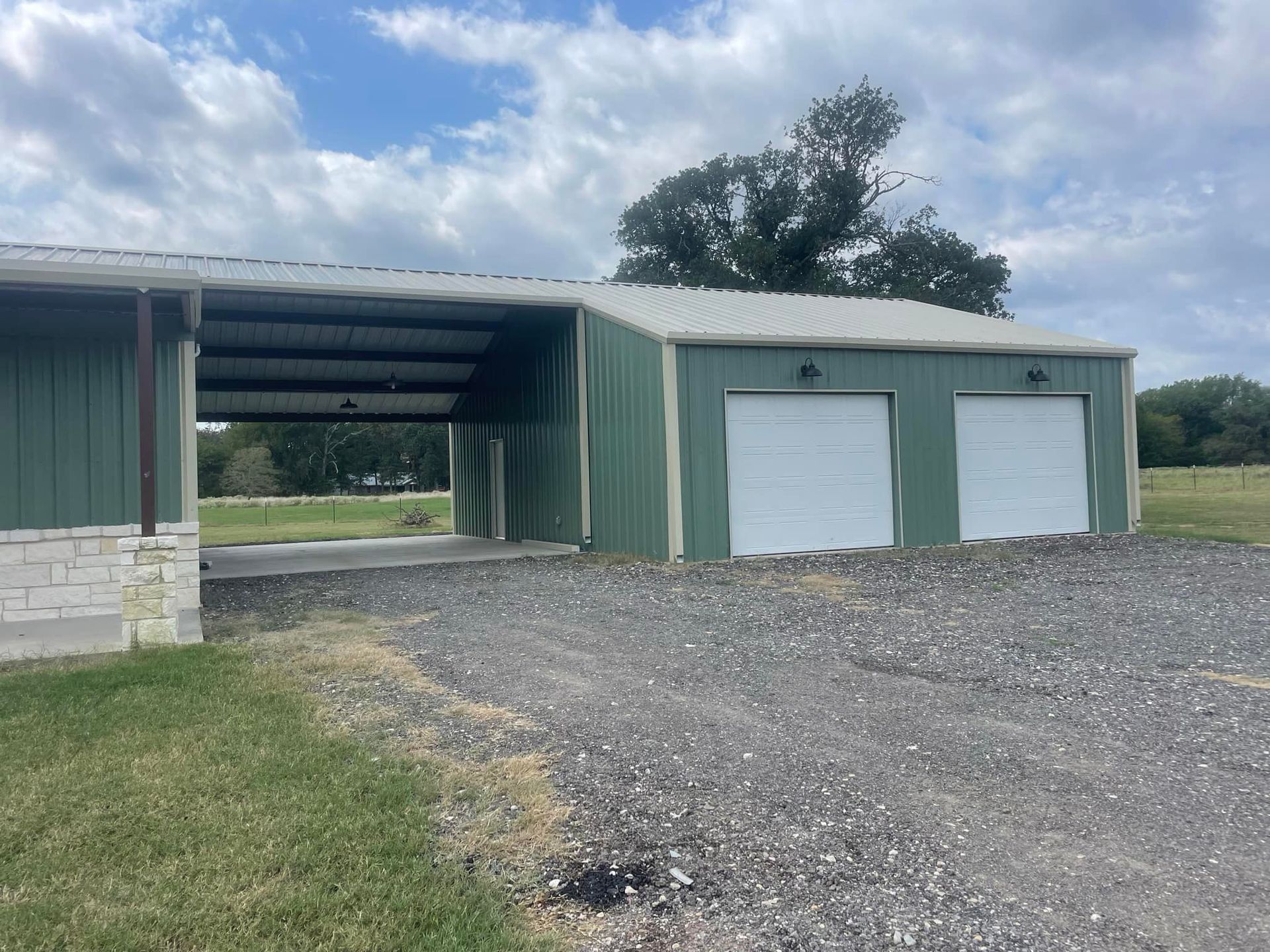 A green garage with two white garage doors is sitting on top of a gravel road.