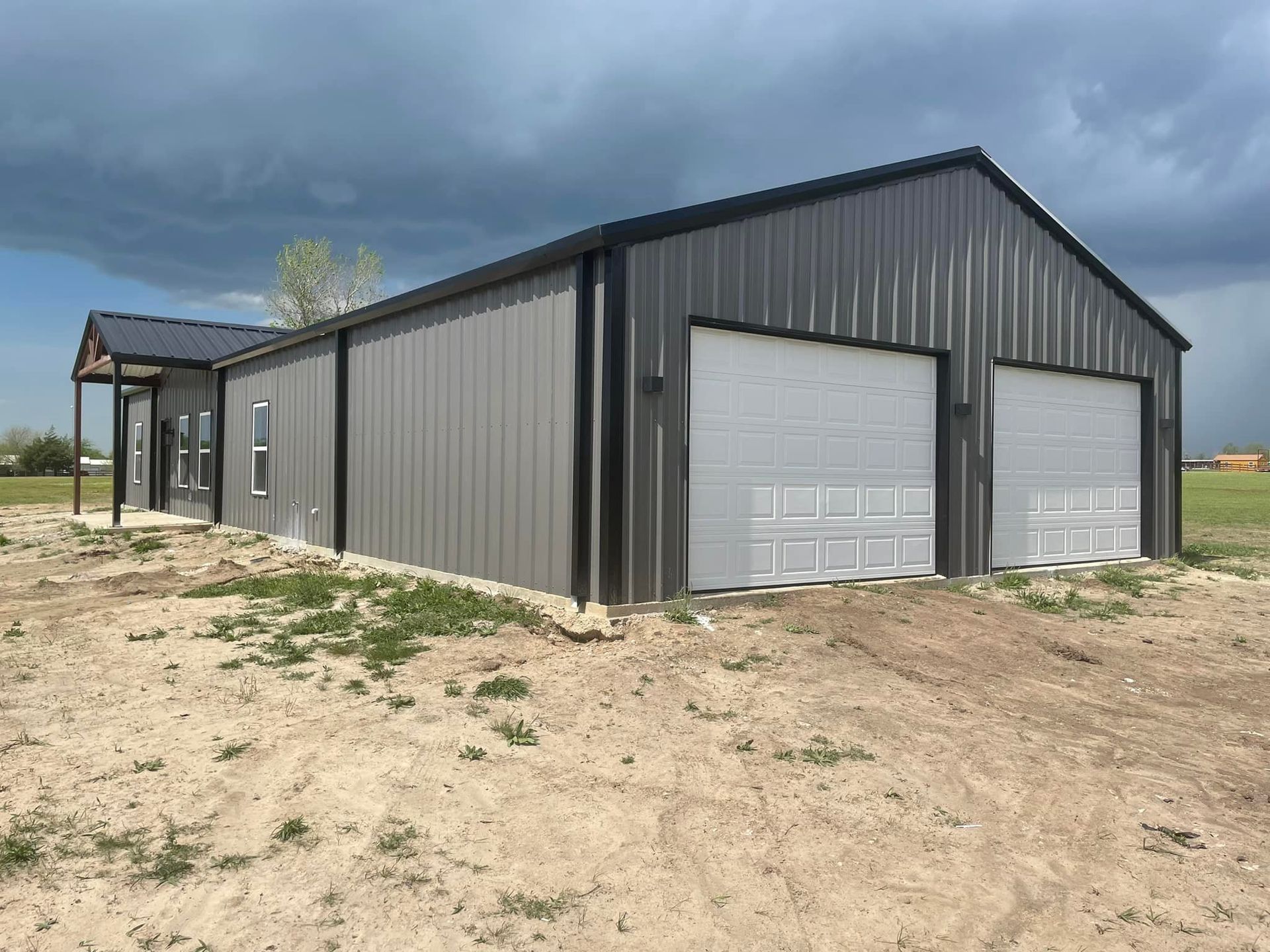 A large metal building with two garage doors is sitting in the middle of a dirt field.