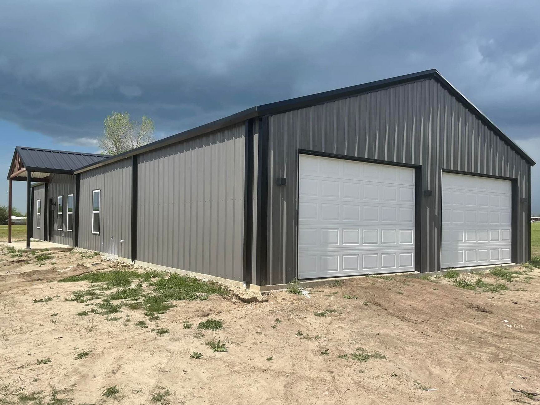 A large metal building with two garage doors is sitting in the middle of a dirt field.