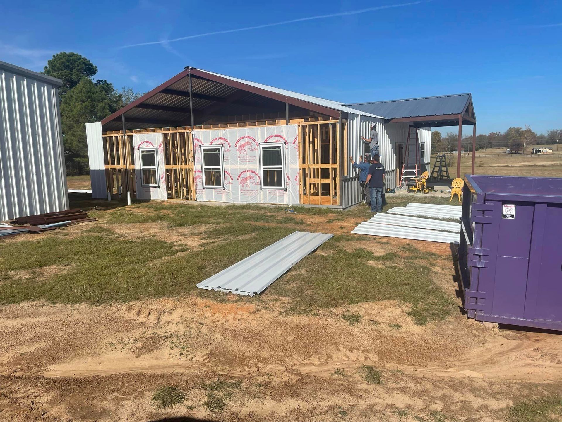 A house is being built in a field next to a purple dumpster.