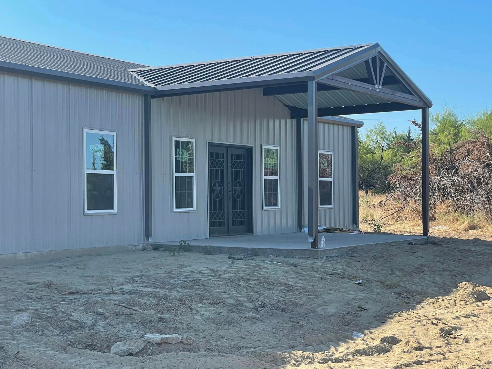 A large metal building with a porch and windows is sitting in the middle of a dirt field.