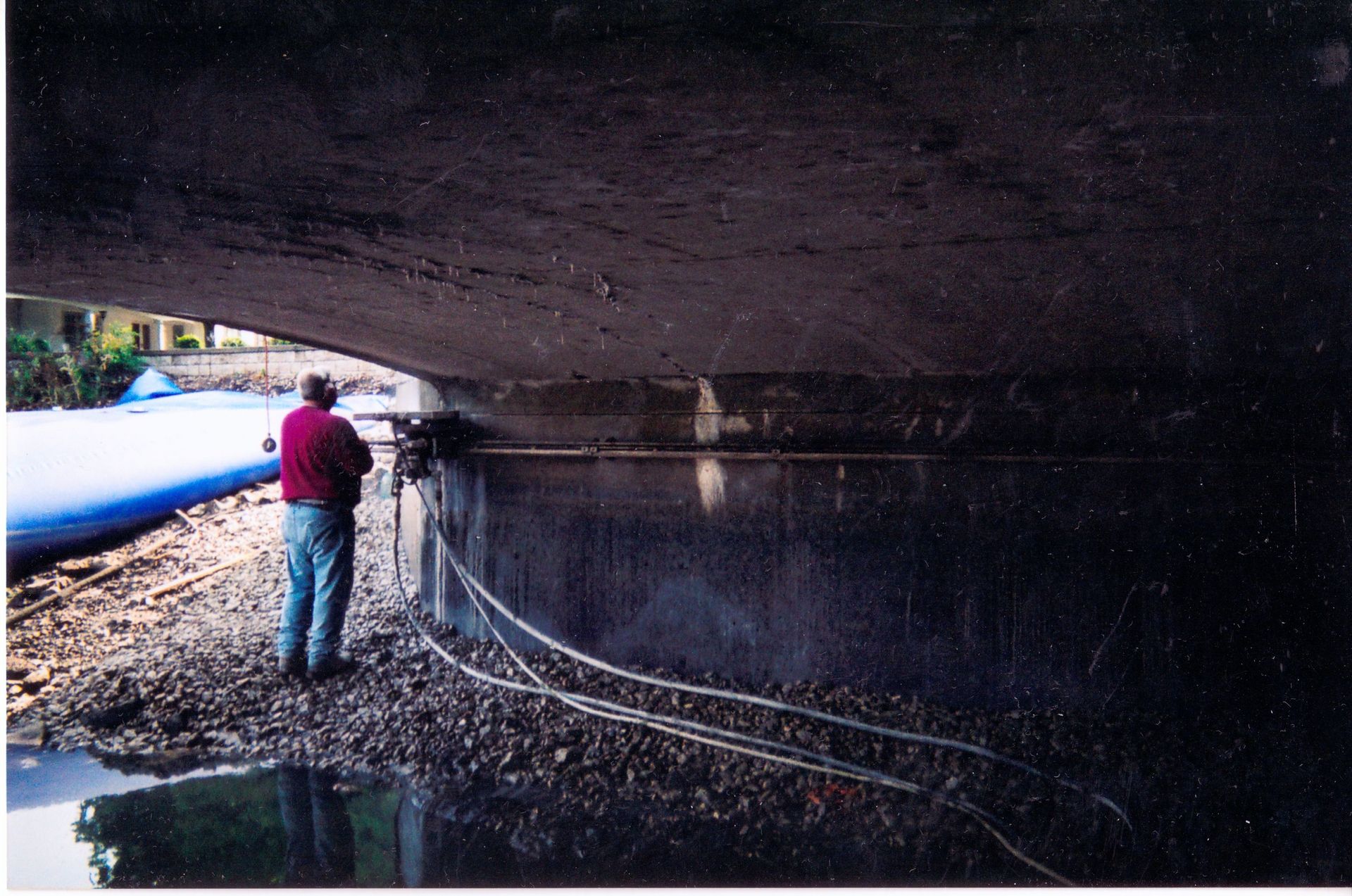 A man in a red jacket stands under a bridge over a body of water