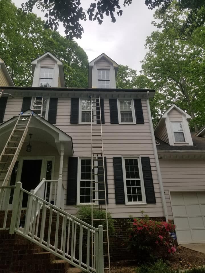 Two-story house with ladders, white siding, black shutters, dormer windows, and trees in the background.