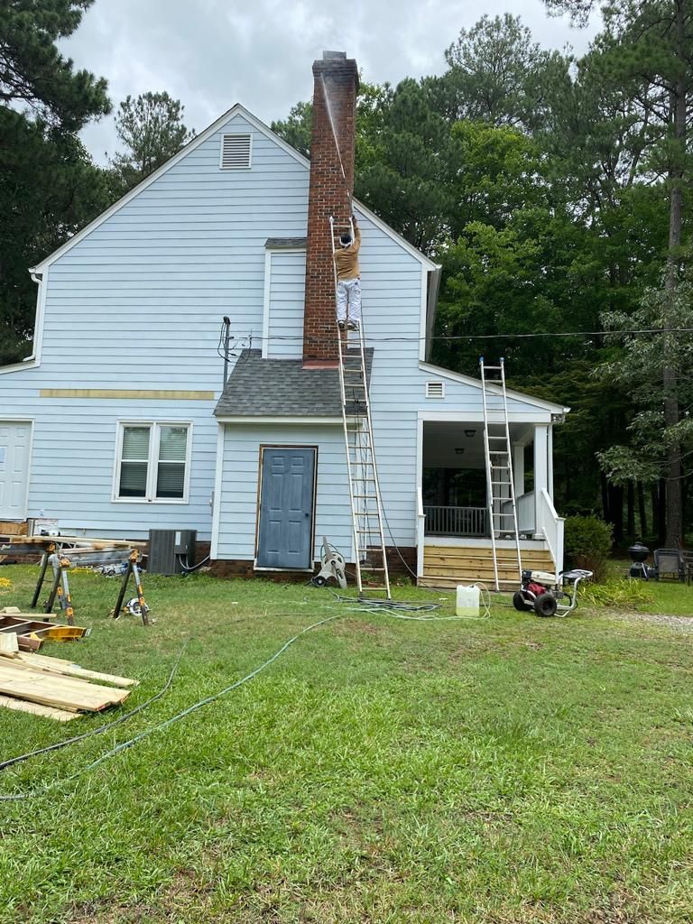 A person on a ladder repairs a tall brick chimney on a blue house. Green grass and trees surround.