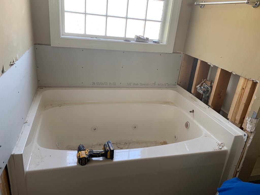 A white jacuzzi tub sits under a window. Construction shows exposed wall studs and drywall. A drill lies on the tub's rim.