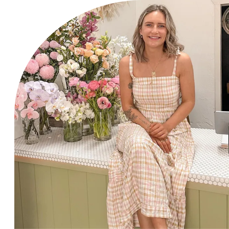 A Woman In A Dress Is Sitting On A Bench In Front Of Vases Of Flowers — Mittagong Flower Merchant in Mittagong, NSW